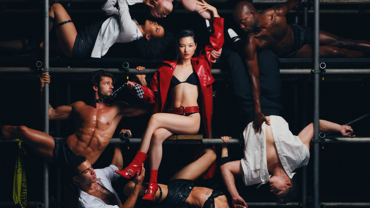 A fashion editorial-style photo featuring Chloe Qisha in a red trench coat, black bikini, and red boots posed on a metal scaffold structure with multiple shirtless models around her.