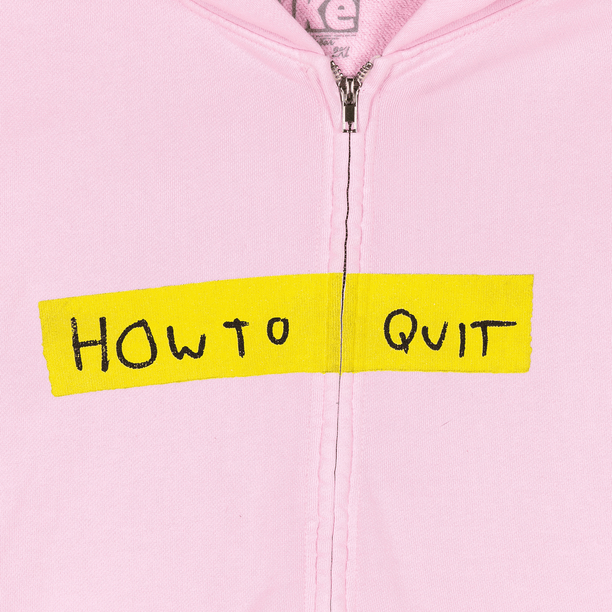 A close-up of a light pink zip-up hoodie with a bright yellow tape strip across the chest reading 'HOW TO QUIT' in black handwritten text.