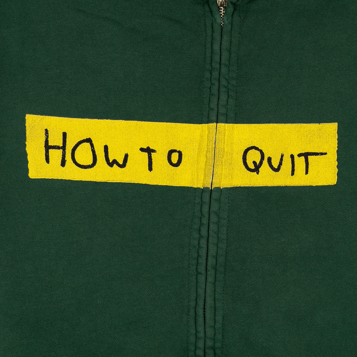 A close-up of a dark green zip-up jacket with a yellow fabric patch across the chest reading 'HOW TO QUIT' in black handwritten-style lettering.
