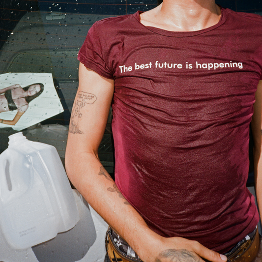 A person wearing a heathered burgundy short-sleeve t-shirt with the white printed text 'The best future is happening' across the chest, photographed in a real environment with a car window, tattoos, and a plastic jug visible.